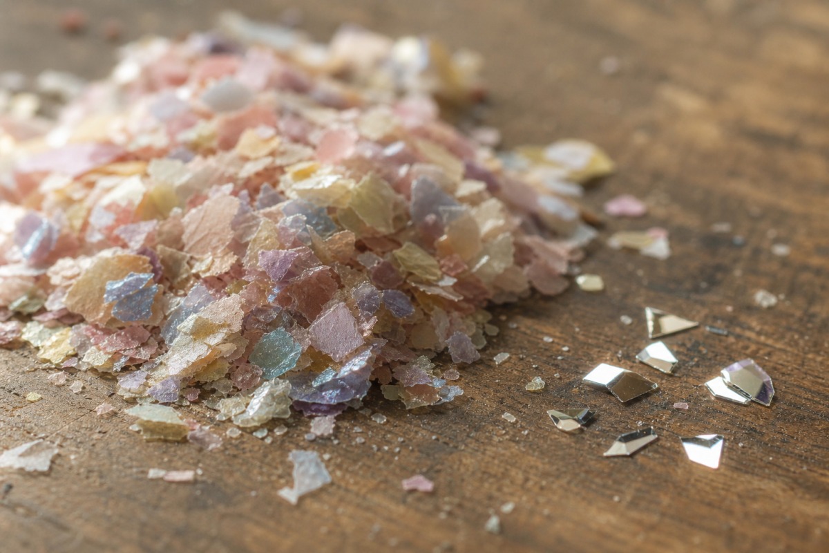 Lähikuva mattapintaisemmasta selluloosapohjaisesta bioglitter-kasasta muutaman kiiltävän synteettisen PET-hiukkasen vieressä puupinnalla.
