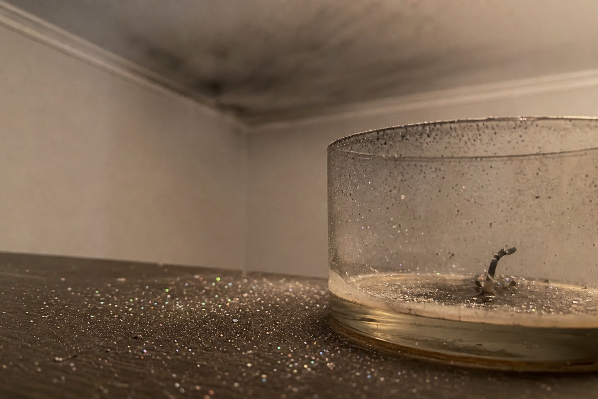 Pieniä kimalluksia ripoteltuna pöydälle loppuun palaneen kynttilän ympärille, taustalla ohut harmaa nokikalvo katossa.